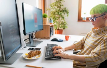 ICT-alantekijä istuu työpöydän ääressä kahden tietokonenäytön edessä ja koodaa.